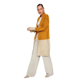 On voit une jeune femme brune sur fond blanc qui porte une gilet bicolore : orange en haut et beige en bas, avec un pantalon blanc ample.