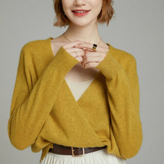 On voit une femme qui porte une gilet cache-coeur jaune avec une jupe plissée blanche et une ceinture, elle touche son pendentif.