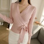 On voit un gilet cache coeur rose sur une femme qui porte les cheveux longs bruns et un pantalon blanc.