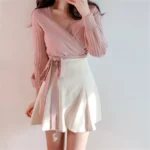 On voit une femme brune qui porte un gilet cache-cœur rose et une jupe blanche.
