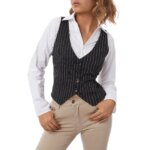 On voit une femme blonde qui porte une chemise blanche, un jean beige et un gilet de costume court noir rayé blanc. Elle a les mains dans les passants du jean et prend la pose.