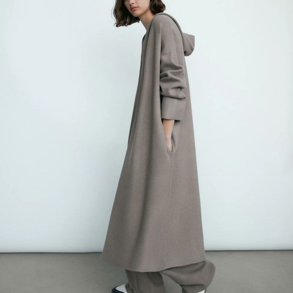 On voit une femme blanche avec un carré brun qui porte un gilet long taupe, marron, avec une capuche. elle marche devant un mur blanc.