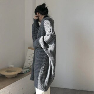 On voit une femme de trois quarts qui regarde de la décoration sur un muret bas. Elle porte un gilet torsadé gris et un pantalon blanc.