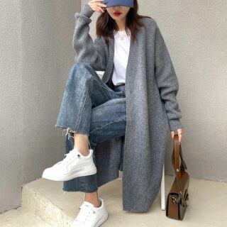 On voit une jeune femme brune avec une casquette assise sur un tabouret. elle porte des baskets blanches, un jean et un gilet gris long oversize.