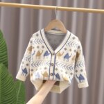 On voit un gilet pour petit garçon beige avec des motifs pastel. Il est suspendu à une trignle.