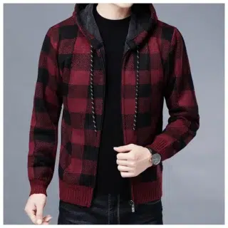 Photo d'un homme debout portant un gilet à capuche à carreauc noirs et blancs sur un pull noiu col haut les mains dans les poches d'un pantalon noir