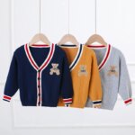 On voit trois gilets pour enfants suspendus à une tringle devant un mur blanc. Les trois gilets sont bleu marine, jaune et gris. Ils ont un écusson ours.