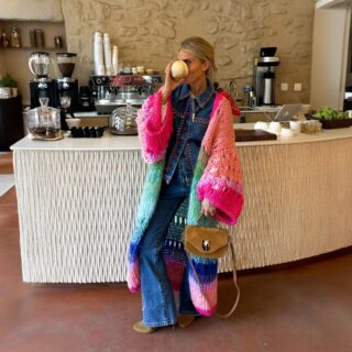 Femme portant un long gilet en maille coloré, avec un deres de couleurs vives, buvant un café.